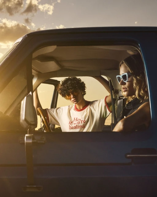 Two women in a blue pickup truck with a sunny sky in the background. One woman drives with yellow sunglasses, the other wears white sunglasses.