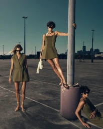 Three models in olive outfits and sunglasses posing on a rooftop parking lot against a skyline view