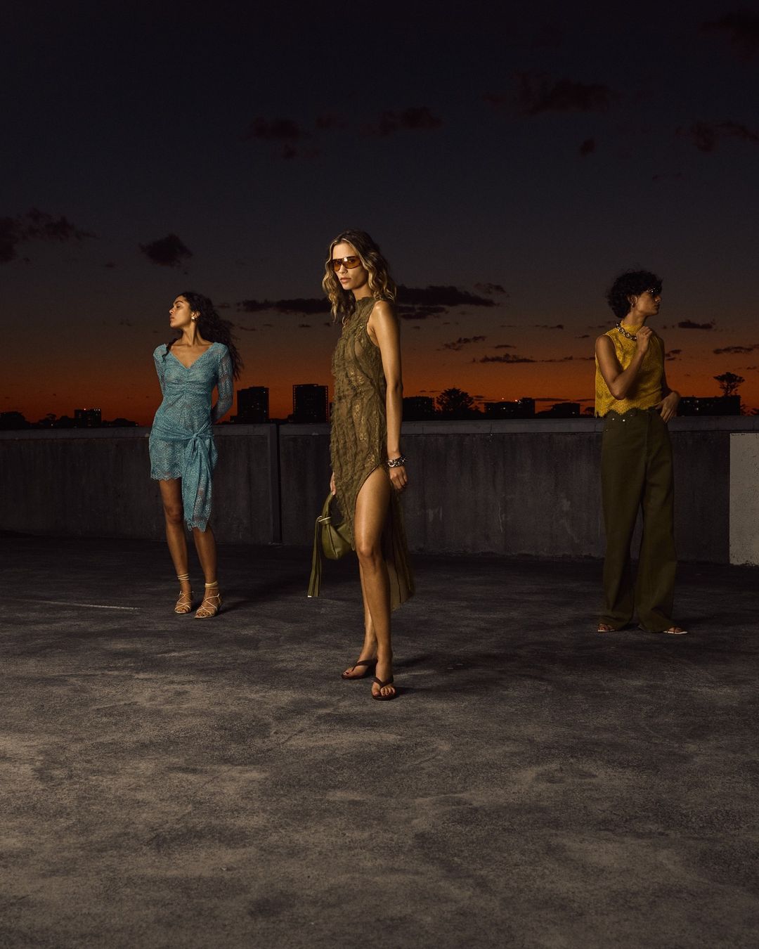 Three models wearing designer fashion stand on a rooftop at dusk with a twilight sky background.
