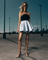 Model wearing a black strapless top and flared white skirt standing on a concrete rooftop against a dark sky.
