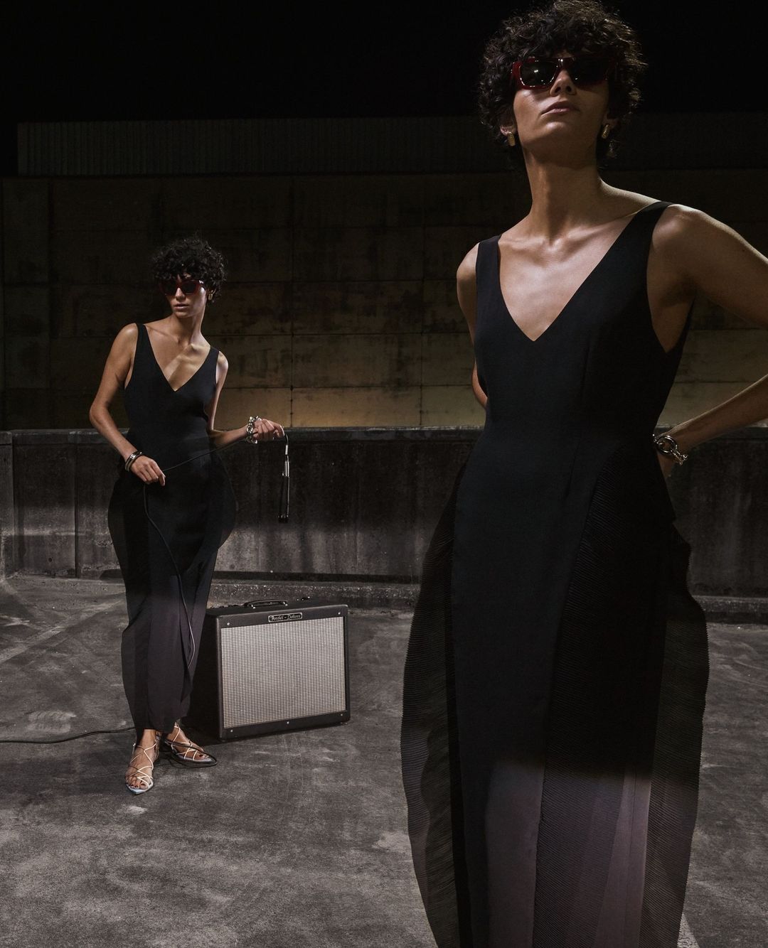 Two women in black dresses and sunglasses pose outdoors with a music amplifier, set against a dark wall, in a fashion editorial.
