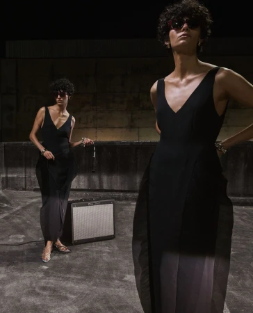 Two women in black dresses and sunglasses pose outdoors with a music amplifier, set against a dark wall, in a fashion editorial.