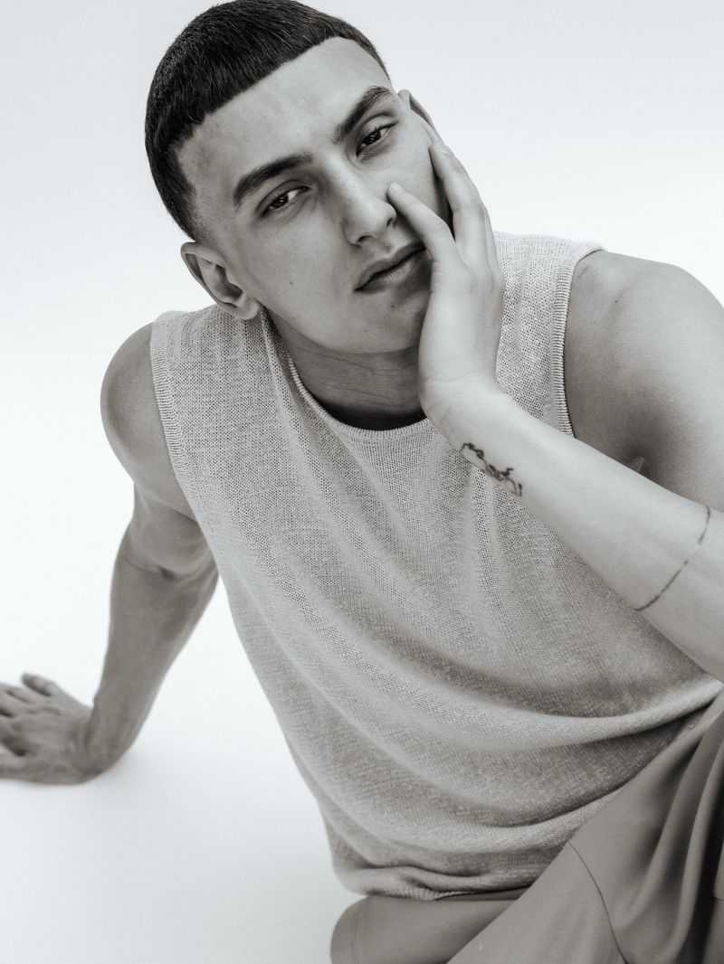 Monochrome portrait of a young adult male with hand to face, wearing a sleeveless top, seated against a plain backdrop in a studio.