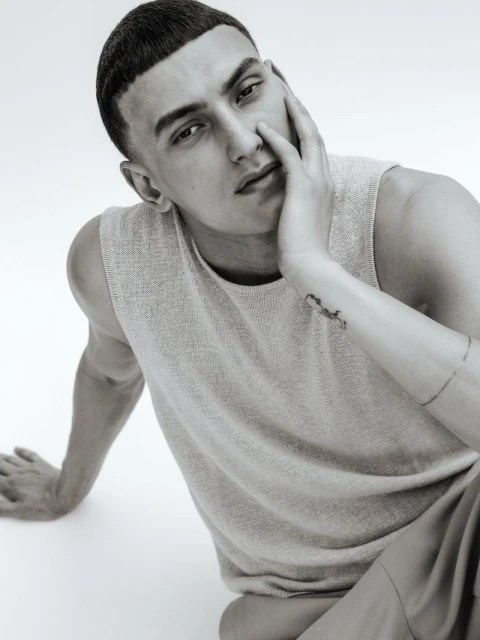 Monochrome portrait of a young adult male with hand to face, wearing a sleeveless top, seated against a plain backdrop in a studio.