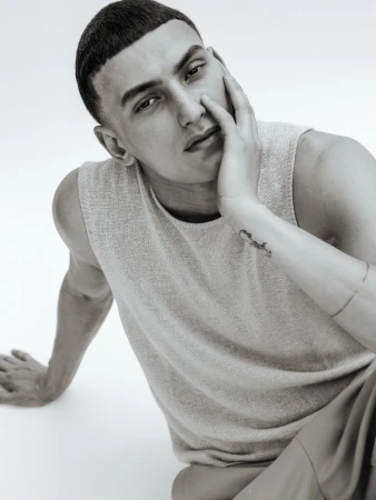 Monochrome portrait of a young adult male with hand to face, wearing a sleeveless top, seated against a plain backdrop in a studio.