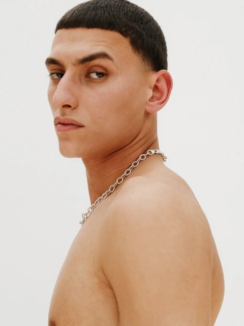 Close-up profile of a young adult man with short dark hair, wearing a silver chain necklace, against a plain white background.