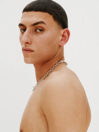 Close-up profile of a young adult man with short dark hair, wearing a silver chain necklace, against a plain white background.