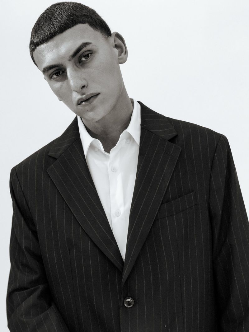 Black and white portrait of a young adult male with a distinctive bowl cut, wearing a pinstripe suit and a white collared shirt against a plain background.
