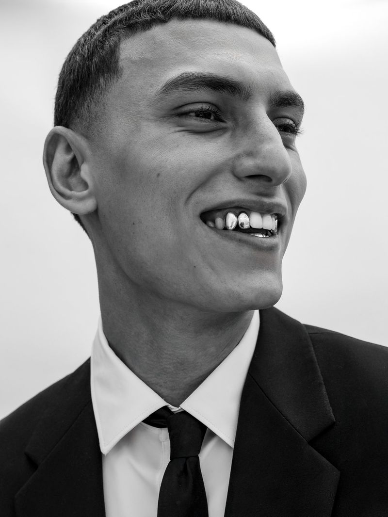 Close-up of a smiling young adult man in a suit and tie, showcasing gold teeth grills. The image is in black and white.