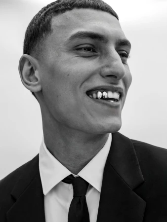Close-up of a smiling young adult man in a suit and tie, showcasing gold teeth grills. The image is in black and white.