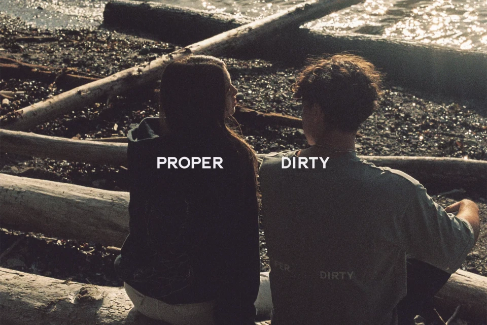 Two young adults sit by the water near logs, wearing casual attire, under sunlight, showing the back of their tops with text.