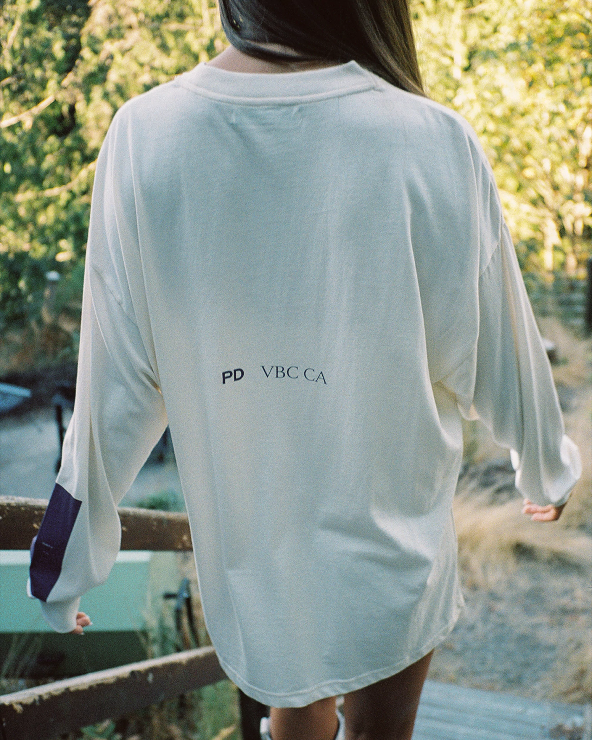 Woman in an oversized long-sleeve t-shirt with 'PD VBCCA' text detail stands outside on stairs with a green foliage backdrop.
