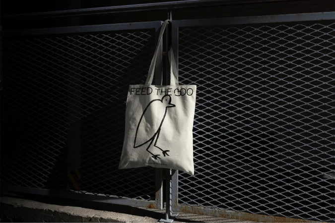 Off-white tote bag with an illustration of a bird and the text 'FEED THE COO' hangs against a dark metal lattice fence.