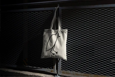 Cream canvas tote bag with a black bird graphic hanging on a dark metal mesh fence under harsh sunlight.