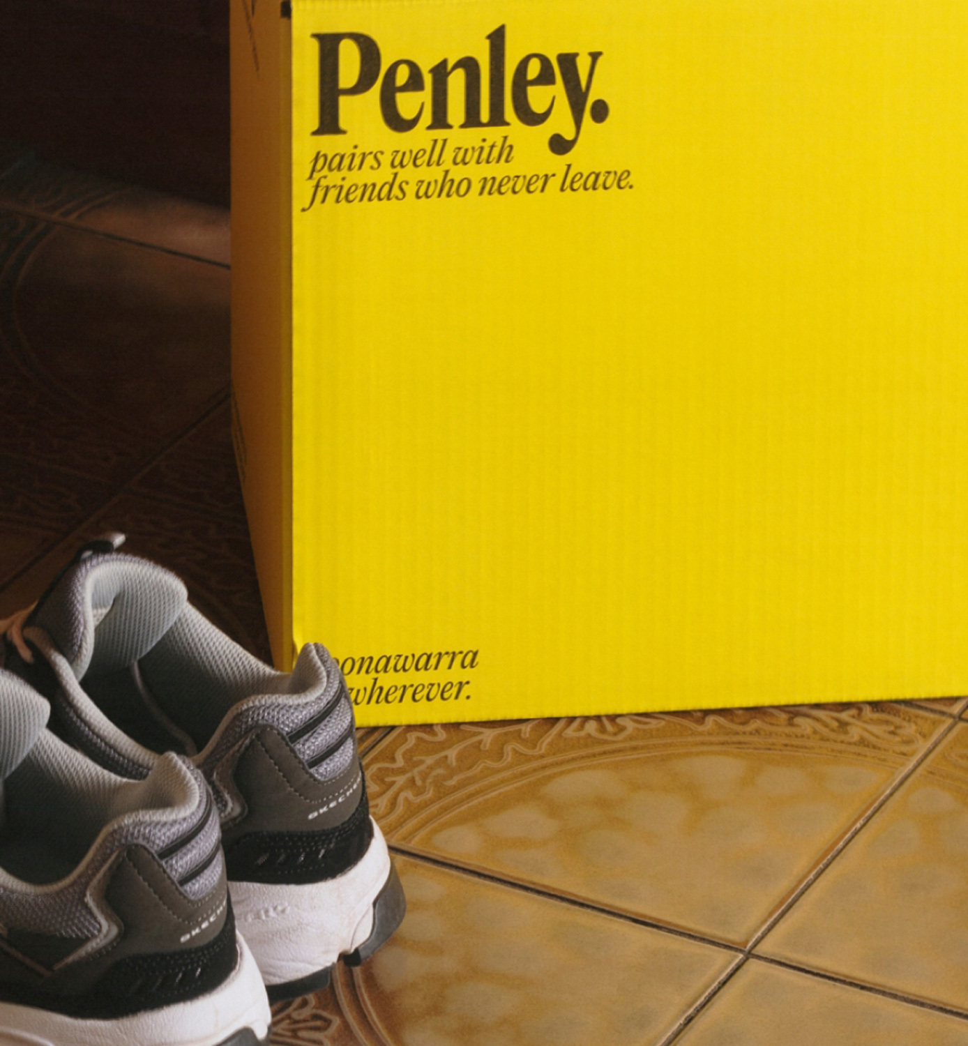 Still life featuring pair of gray and white sneakers on tile floor next to yellow Penley box with advertising text indoors.