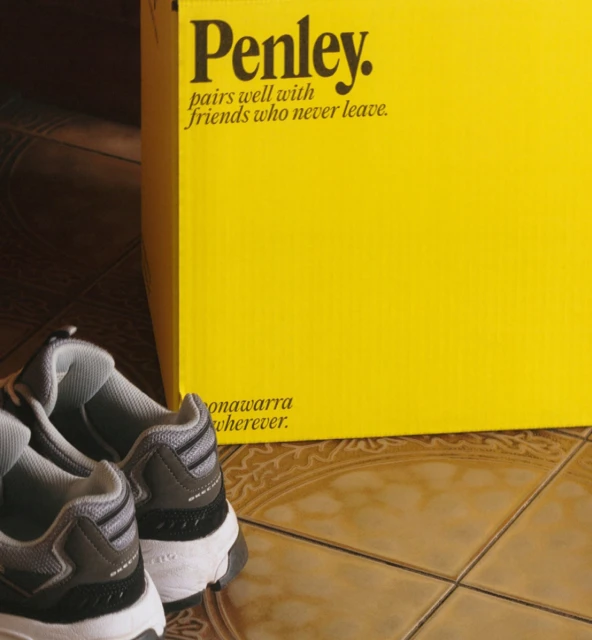 Still life featuring pair of gray and white sneakers on tile floor next to yellow Penley box with advertising text indoors.