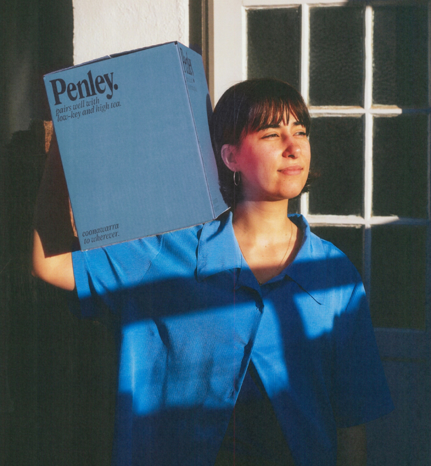 A young adult woman carries a blue box with the brand name Penley on her shoulder in front of a white door and a blue wall.