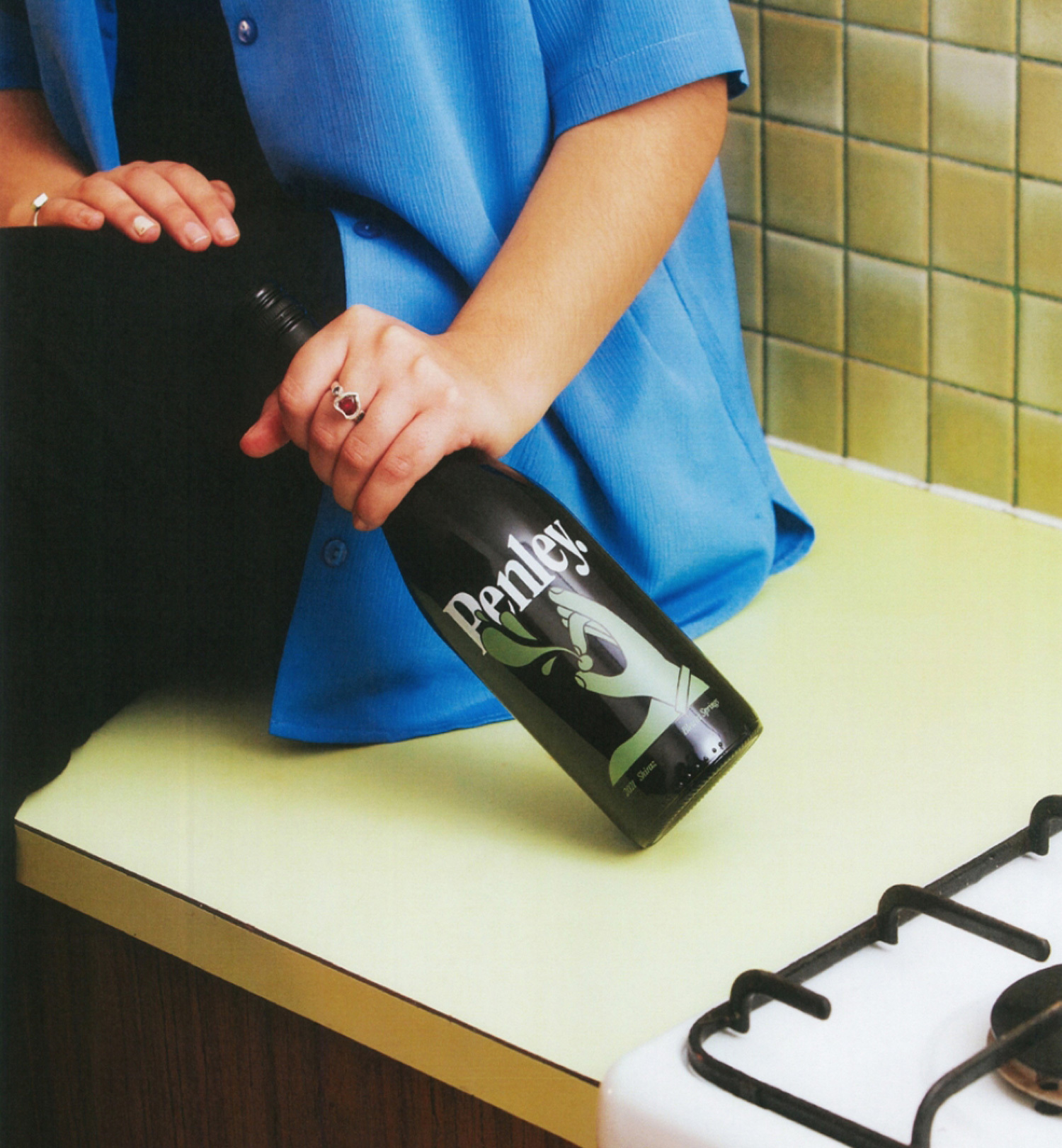 Person in blue top holds Penley bottle on countertop near stove. Kitchen setting with olive green tile and retro aesthetic.