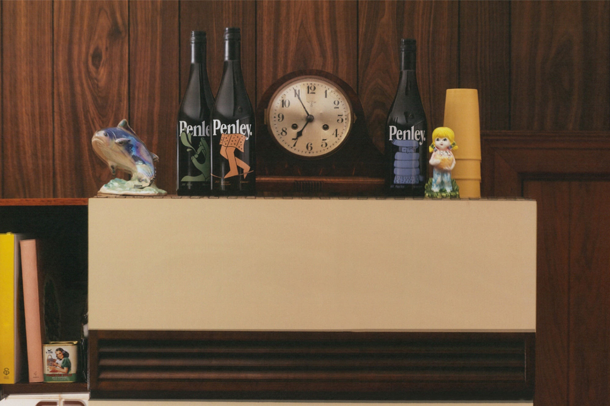 Still life shot of vintage Penley wine bottles alongside retro figurines and an analog clock on a shelf against a wood-paneled wall.