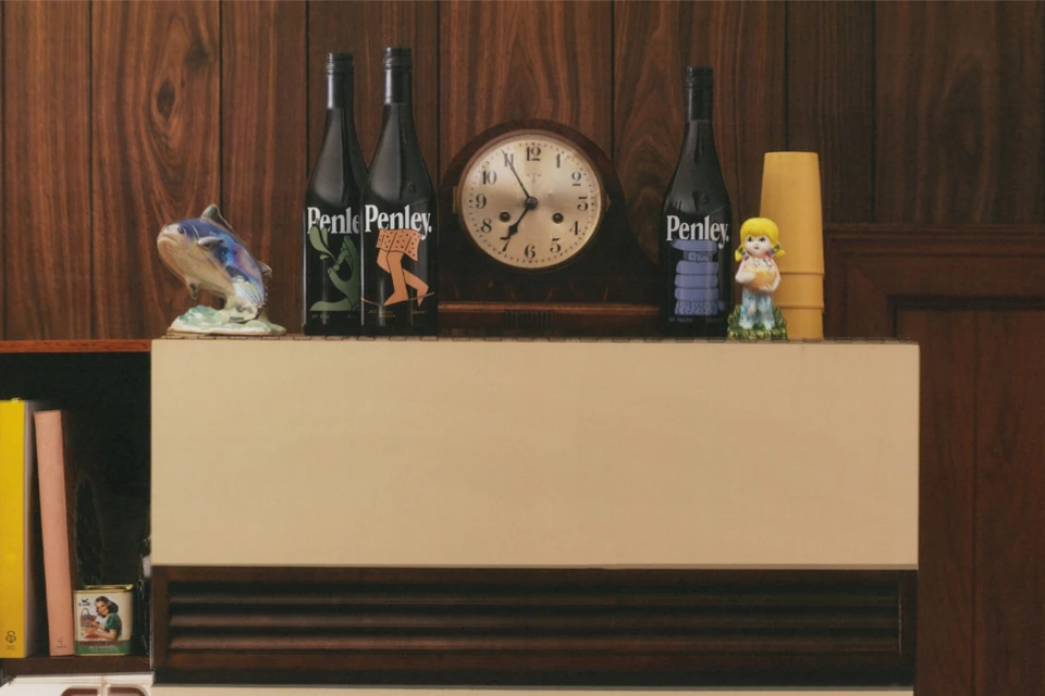 Still life shot of vintage Penley wine bottles alongside retro figurines and an analog clock on a shelf against a wood-paneled wall.