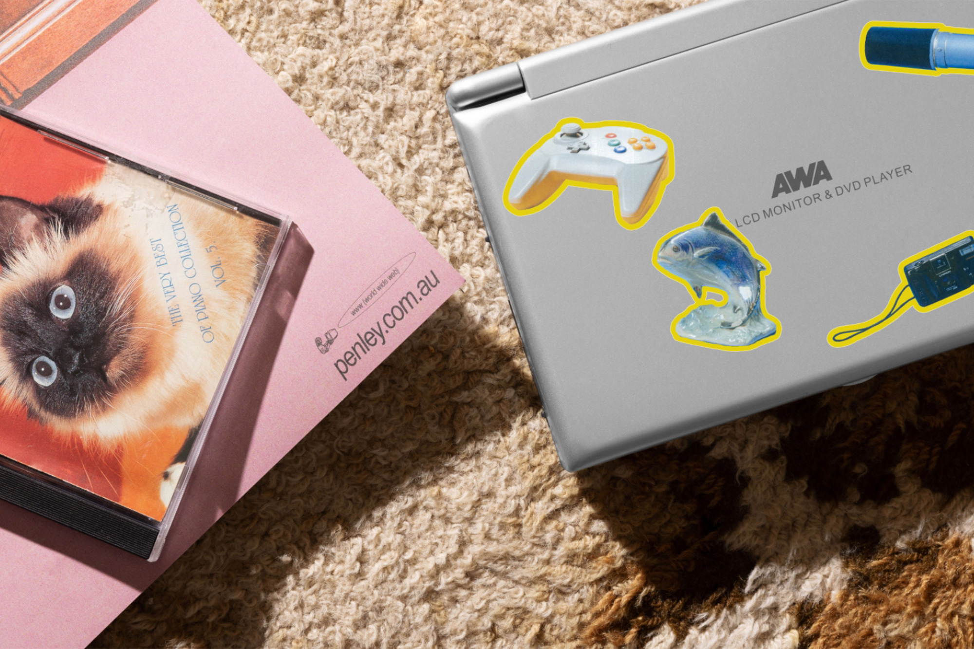 Still life of a laptop adorned with stickers, a pink book, and a CD case featuring a cat, all resting on a textured rug.