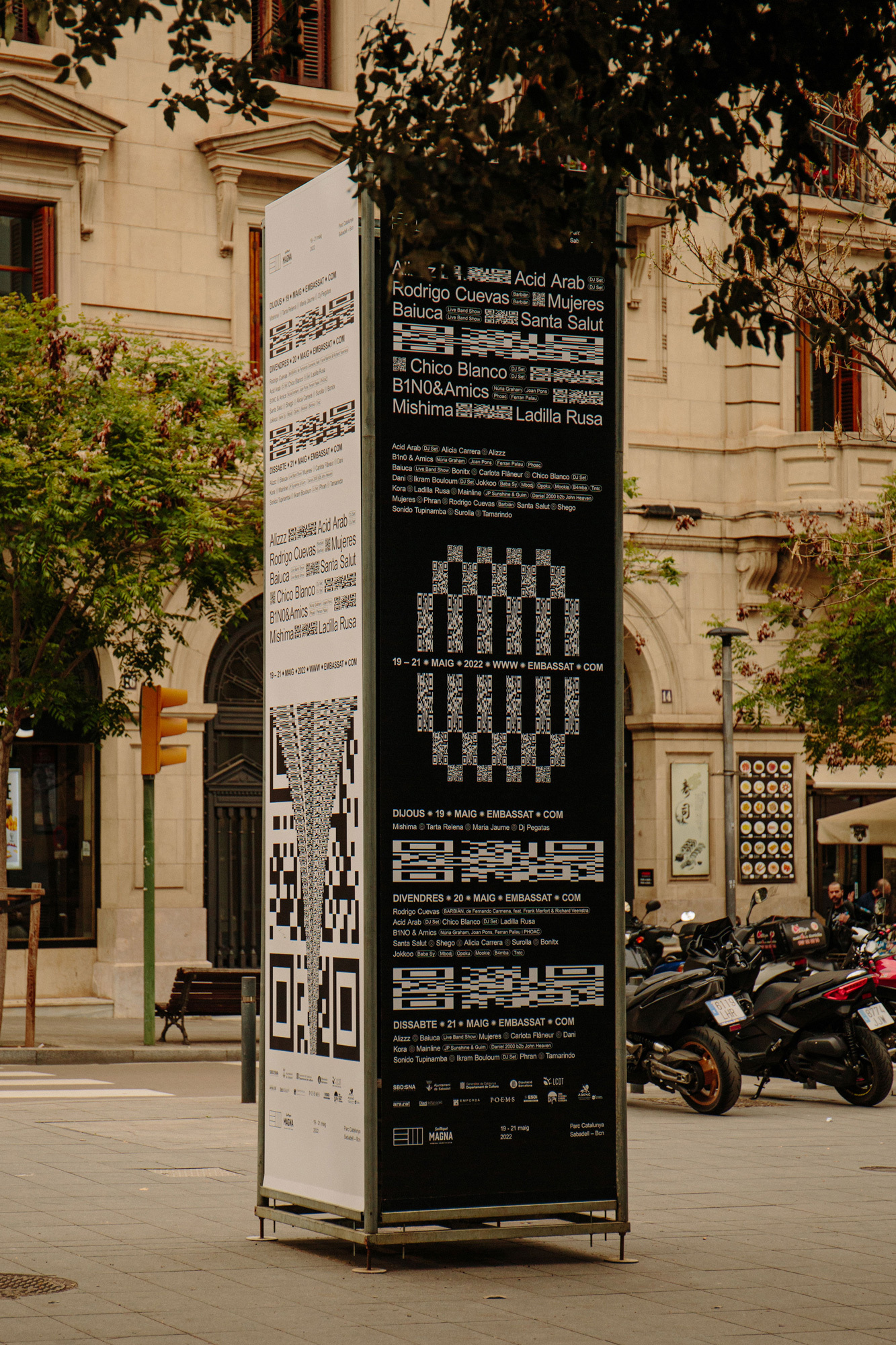 Outdoor poster stand with QR codes and event information in black and white, with architecture and motorcycles in the background.