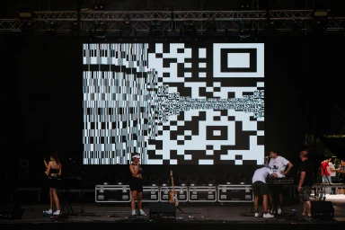Stage performance with musicians below a large digital screen displaying a black and white pixelated graphic design pattern
