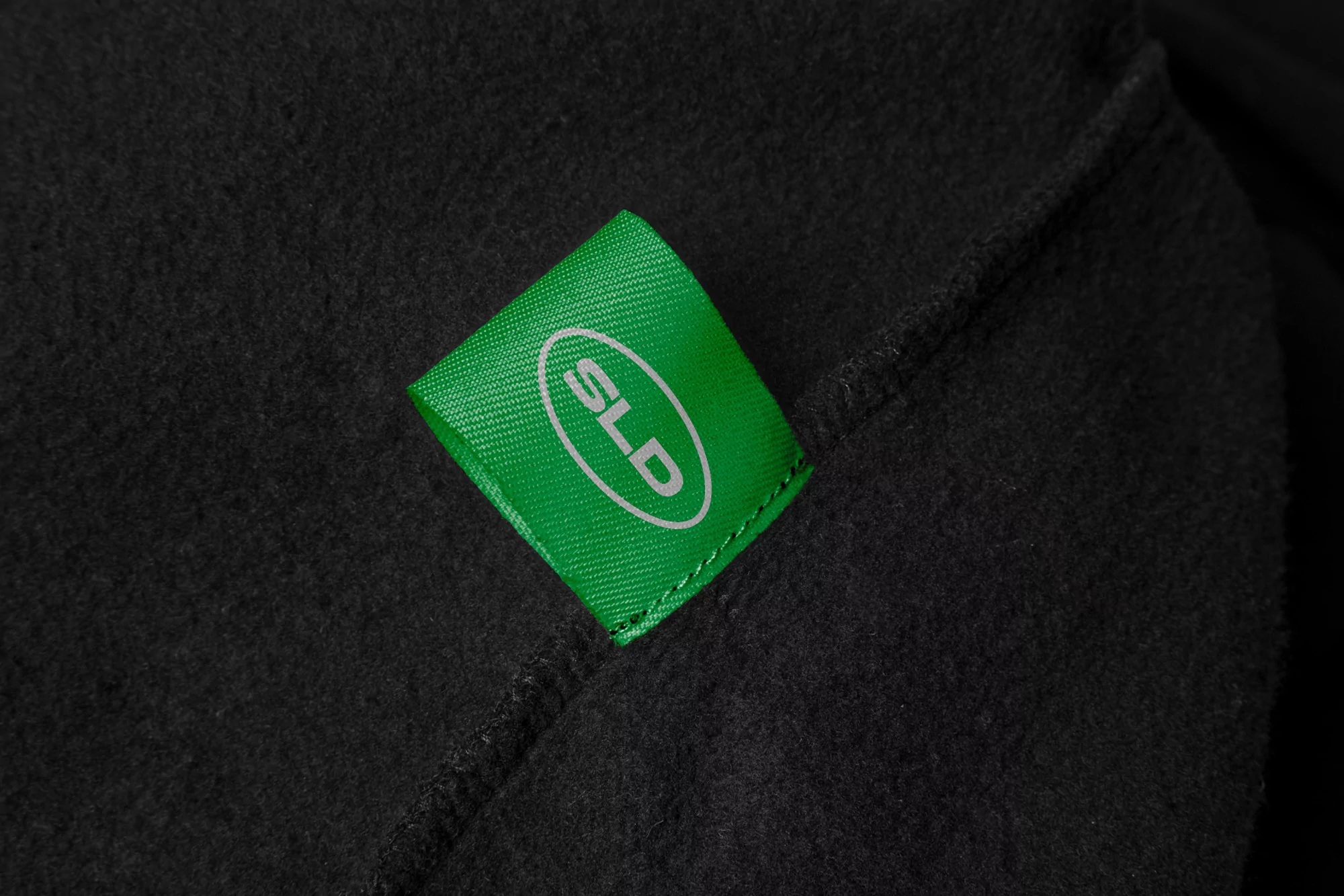 Close-up on a green tag featuring white 'SLD' text, sewn onto a textured black fabric, possibly a garment or accessory.