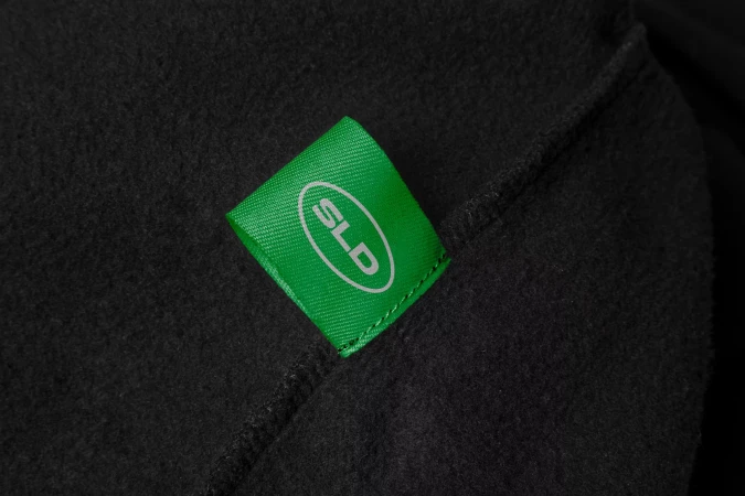 Close-up on a green tag featuring white 'SLD' text, sewn onto a textured black fabric, possibly a garment or accessory.