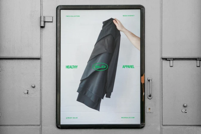 Fashion advertising poster featuring a layered black jacket held by a light-skinned hand against a light grey urban background.