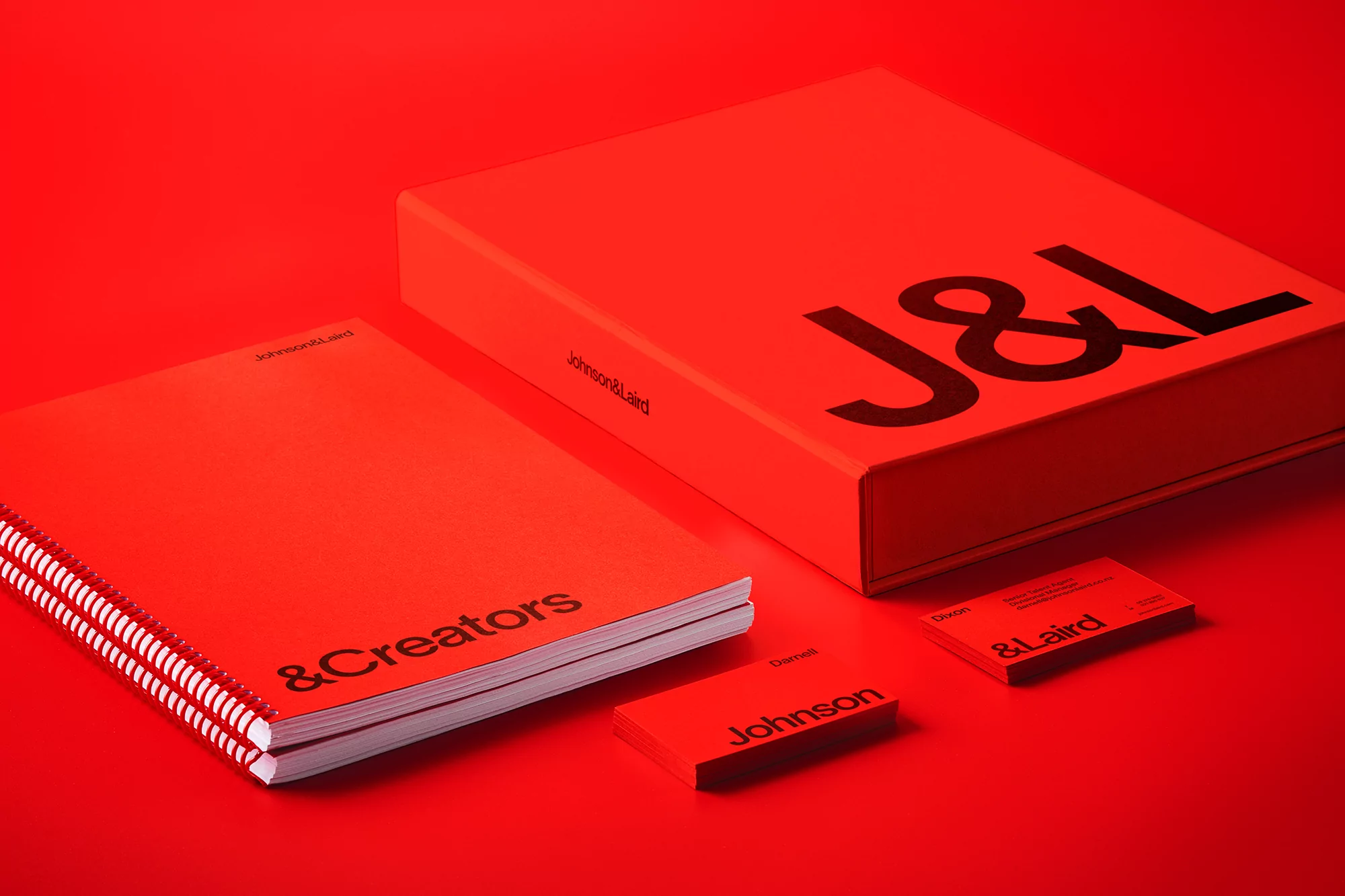 Promotional image featuring a red box with the letters J&L, a notebook with & Creators, and business cards, all set against a red backdrop.