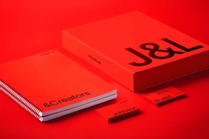Promotional image featuring a red box with the letters J&L, a notebook with & Creators, and business cards, all set against a red backdrop.