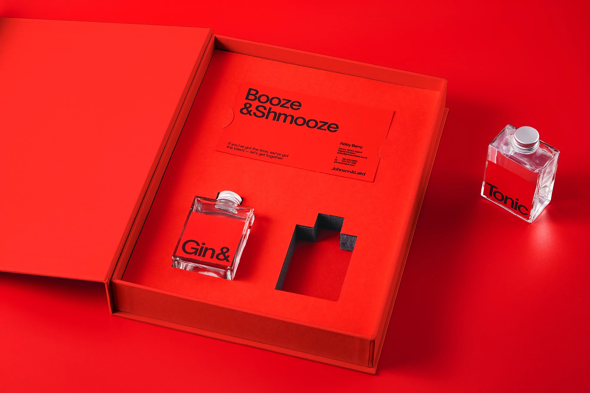 Close-up of an open red box containing a small bottle of gin, with a small bottle of tonic beside it on a matching red surface.