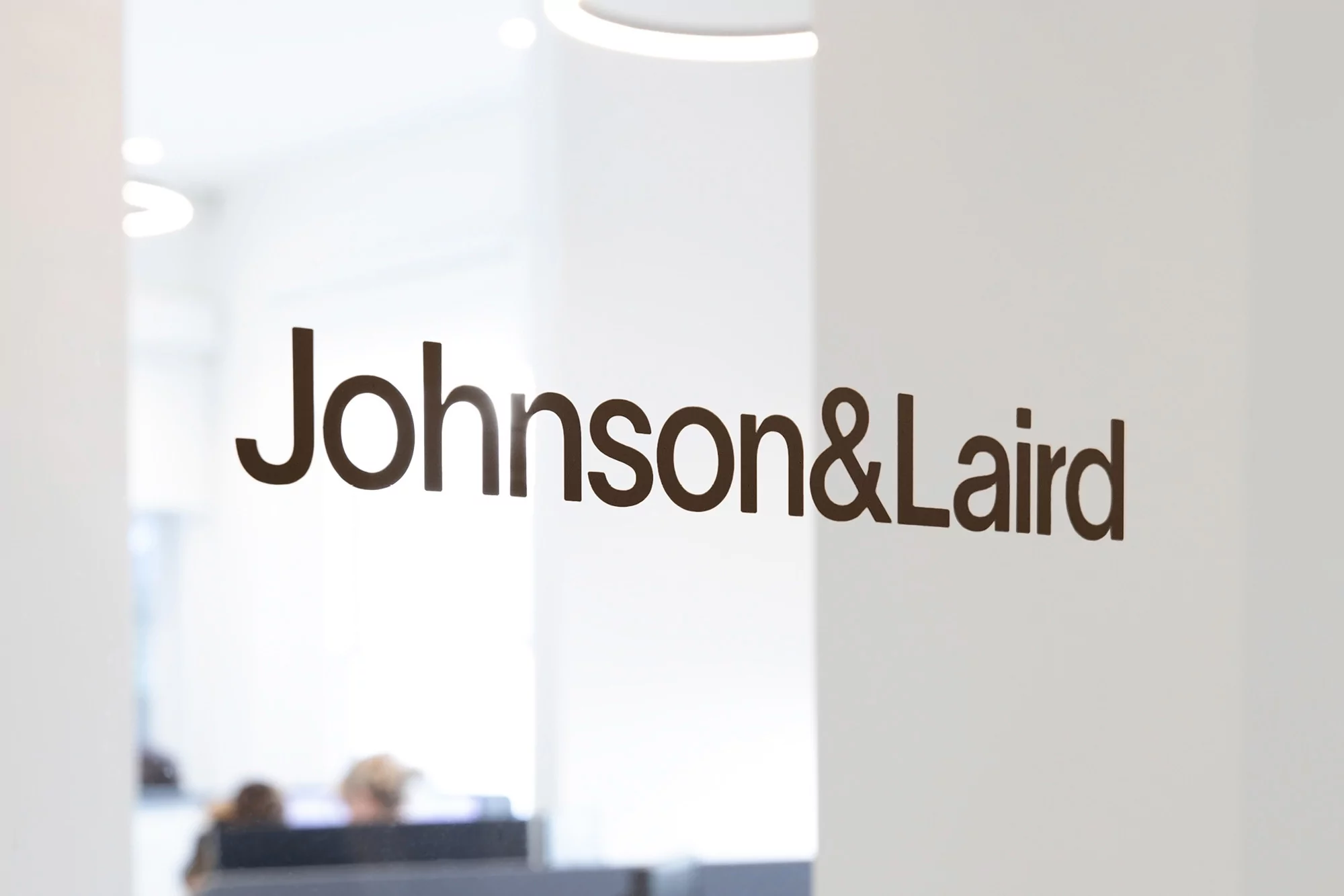 Close-up of the Johnson&Laird company logo on a glass partition inside an office setting, with people visible in the background.