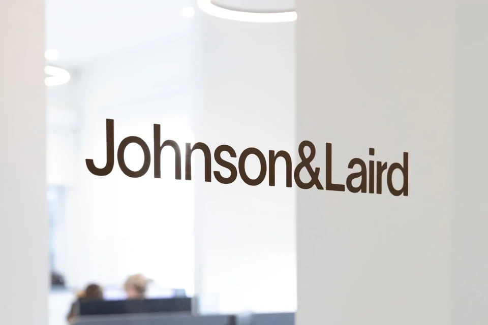 Close-up of the Johnson&Laird company logo on a glass partition inside an office setting, with people visible in the background.