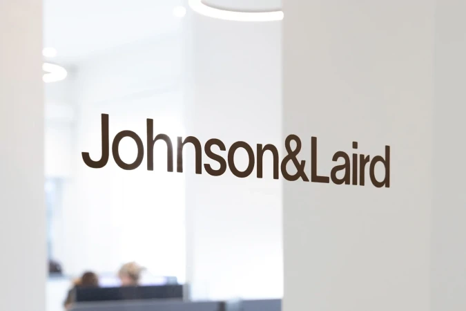 Close-up of the Johnson&Laird company logo on a glass partition inside an office setting, with people visible in the background.