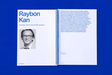 Open spiral-bound brochure on a solid blue background featuring bold blue typography, portrait, and biographical text about Raybon Kan.