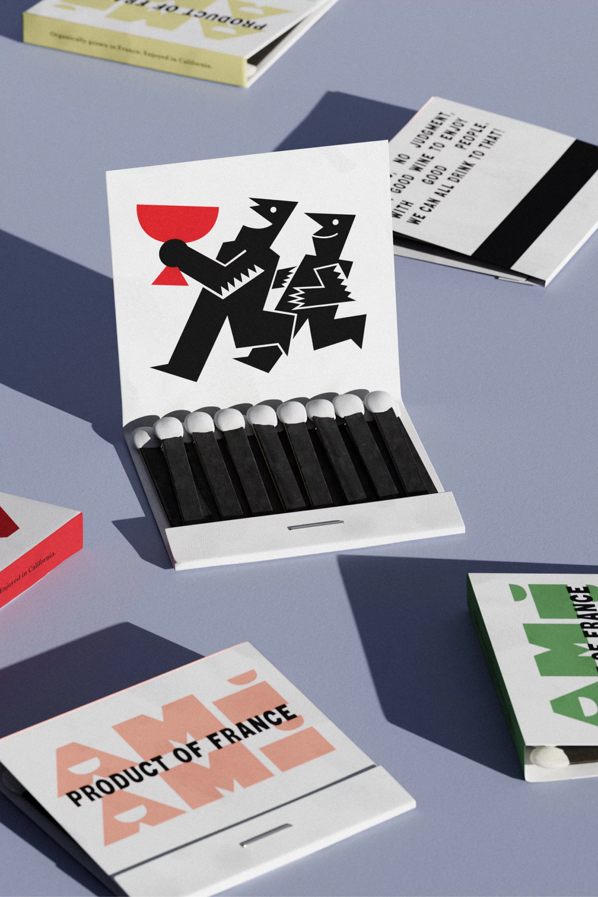 Still life photo of a matchbox with a graphic design of two people carrying a red wine glass, surrounded by other matchboxes on a blue surface.