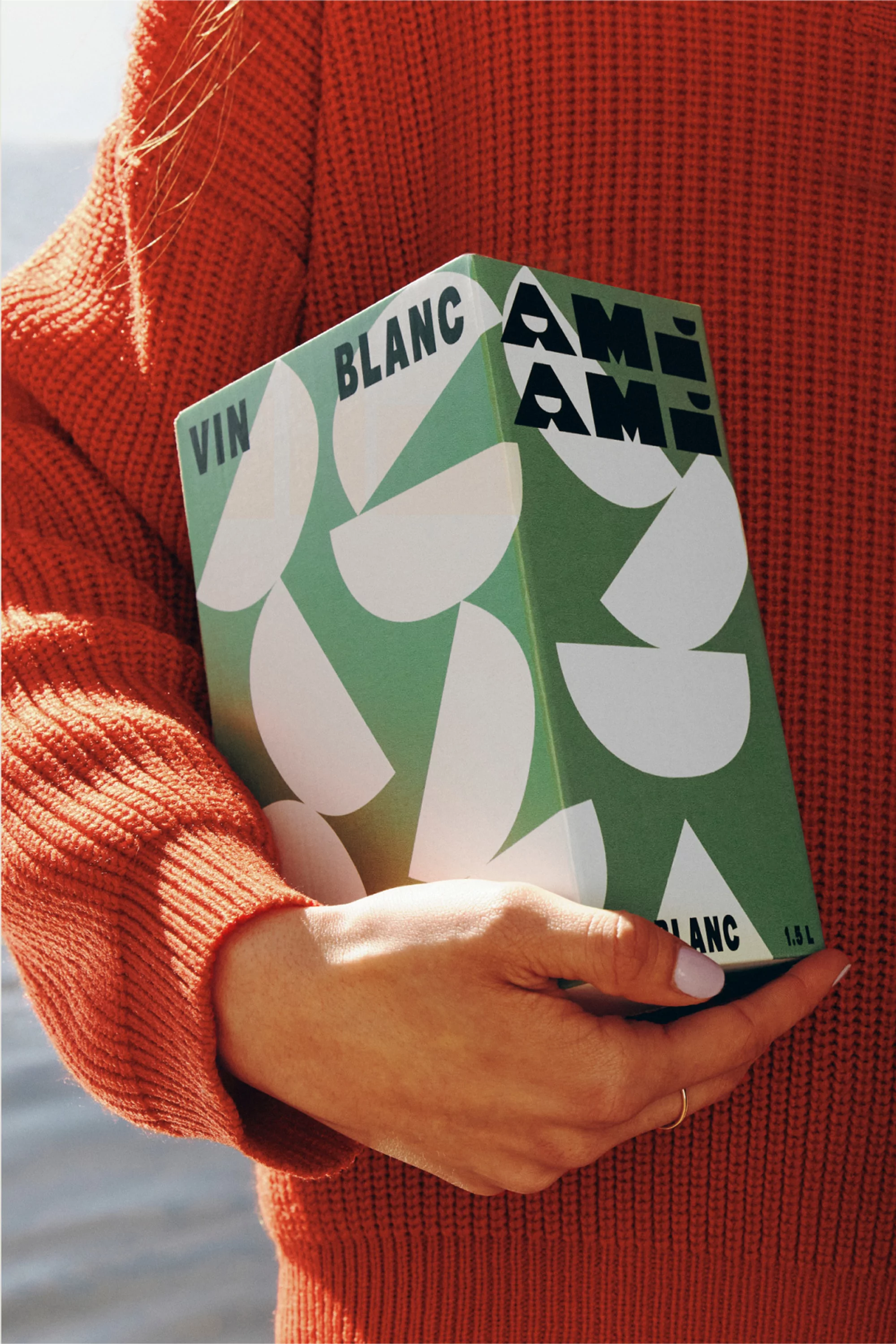 Wine carton held by a woman wearing an orange sweater. The carton is green and white with geometric designs and modern typography.