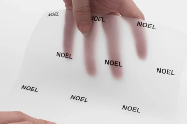 Hands holding a translucent vellum sheet printed with the NOEL brand logo against a clean white background