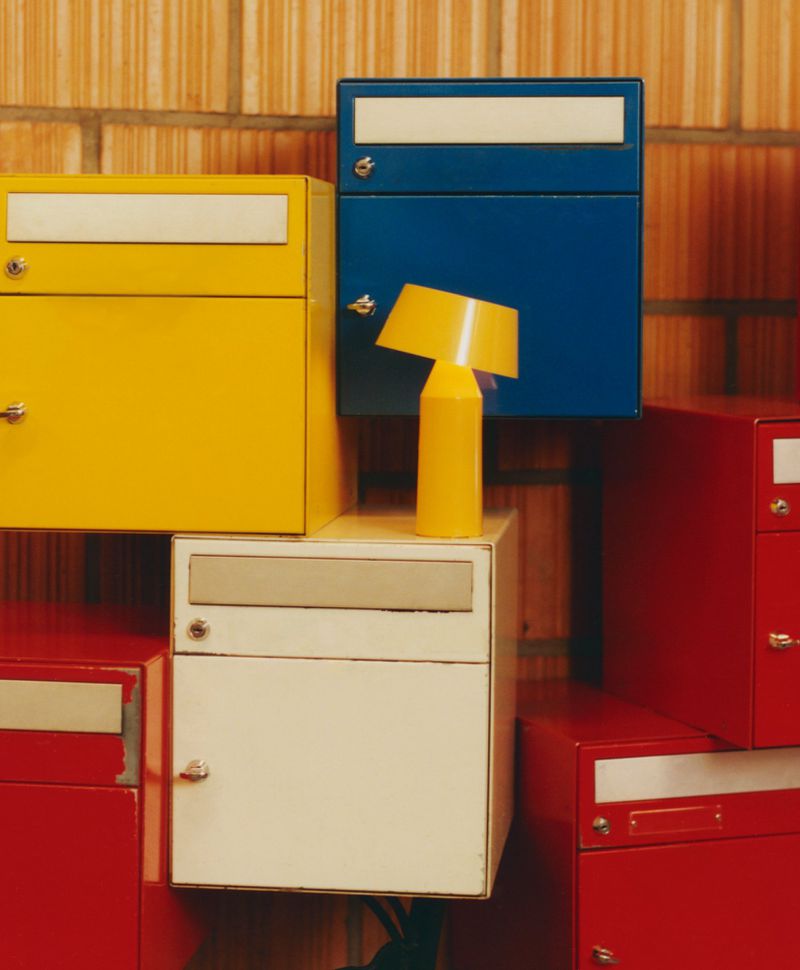 A stack of red, yellow, blue and white metal mailboxes are stacked in front of a beige and tan brick wall with a yellow lamp.