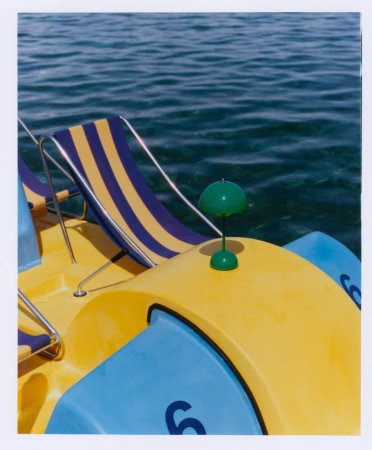 A yellow and blue pedal boat sits on calm, turquoise water. The boat features striped lounge chairs and a bright green accent lamp.