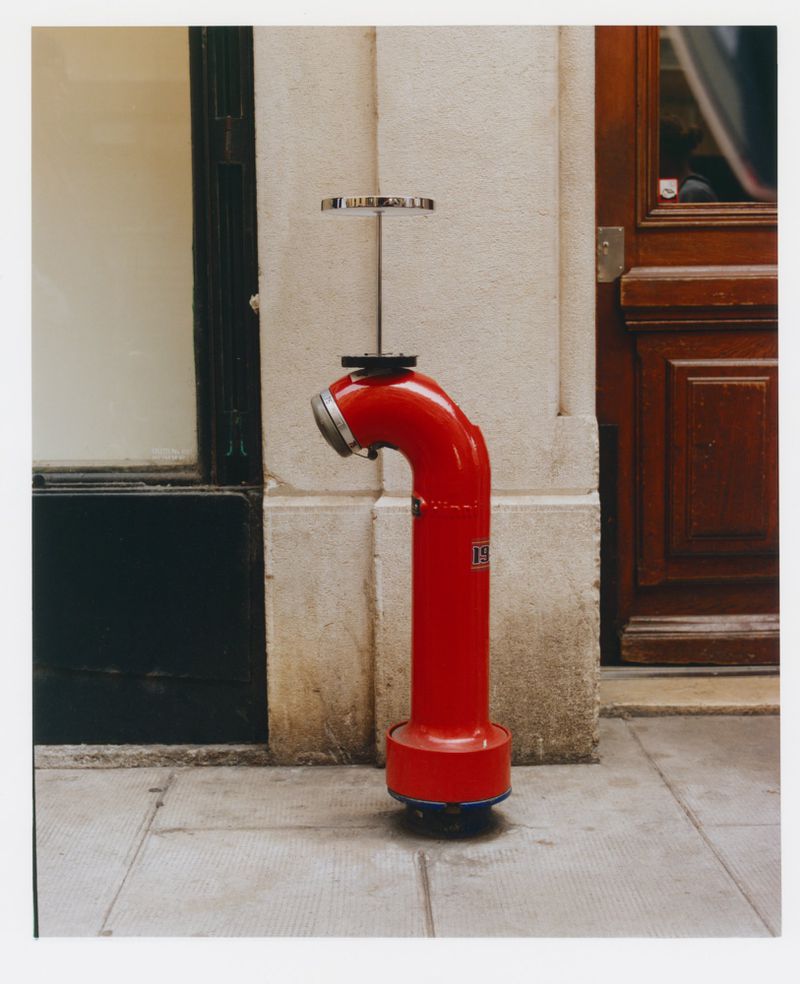 Red fire hydrant stands on a stone sidewalk near building with a black window frame and a wooden door in the background.