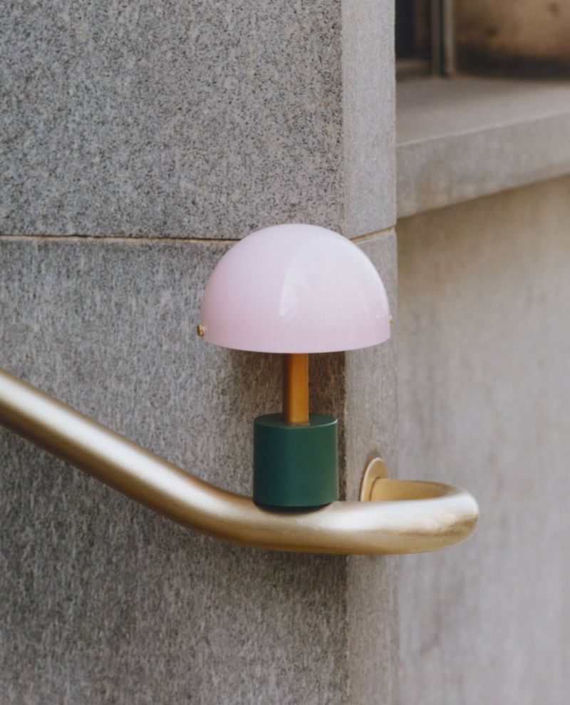 Small, pink mushroom-shaped lamp on a gold railing against a grey stone wall, creating a minimalist, modern, and eye-catching aesthetic.