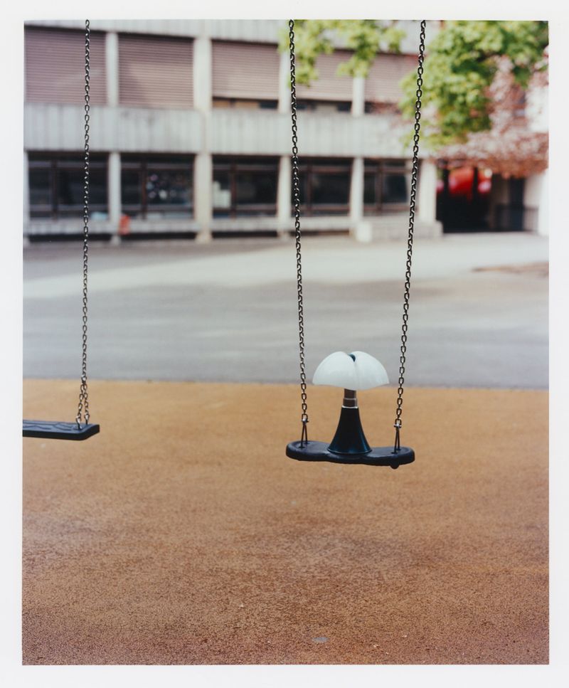 Two swings hang suspended in a playground setting, with a school building and green trees in the background on an overcast day.