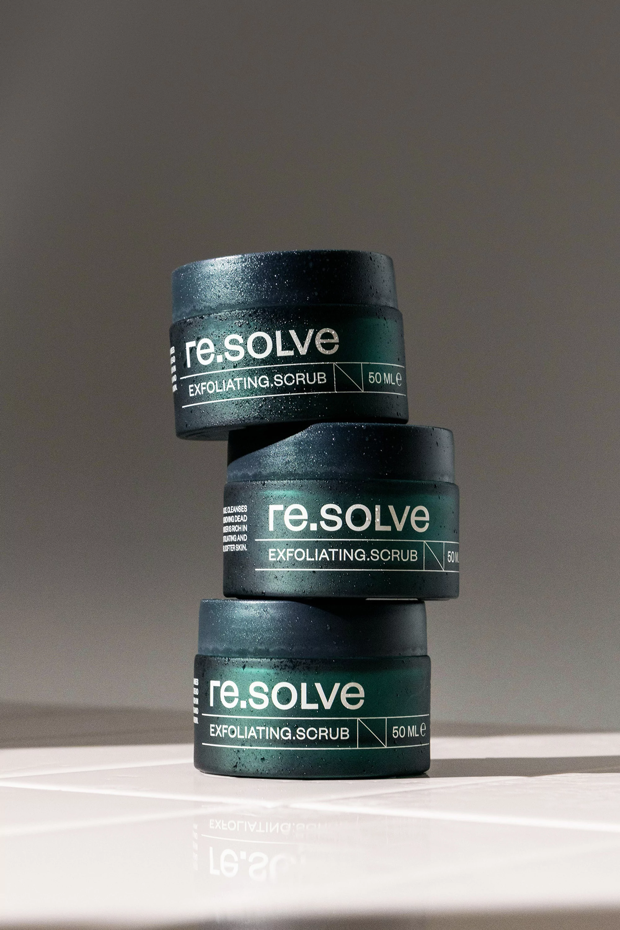 Three stacked jars of re.solve exfoliating scrub on a light surface against a neutral background in a vertical studio shot.