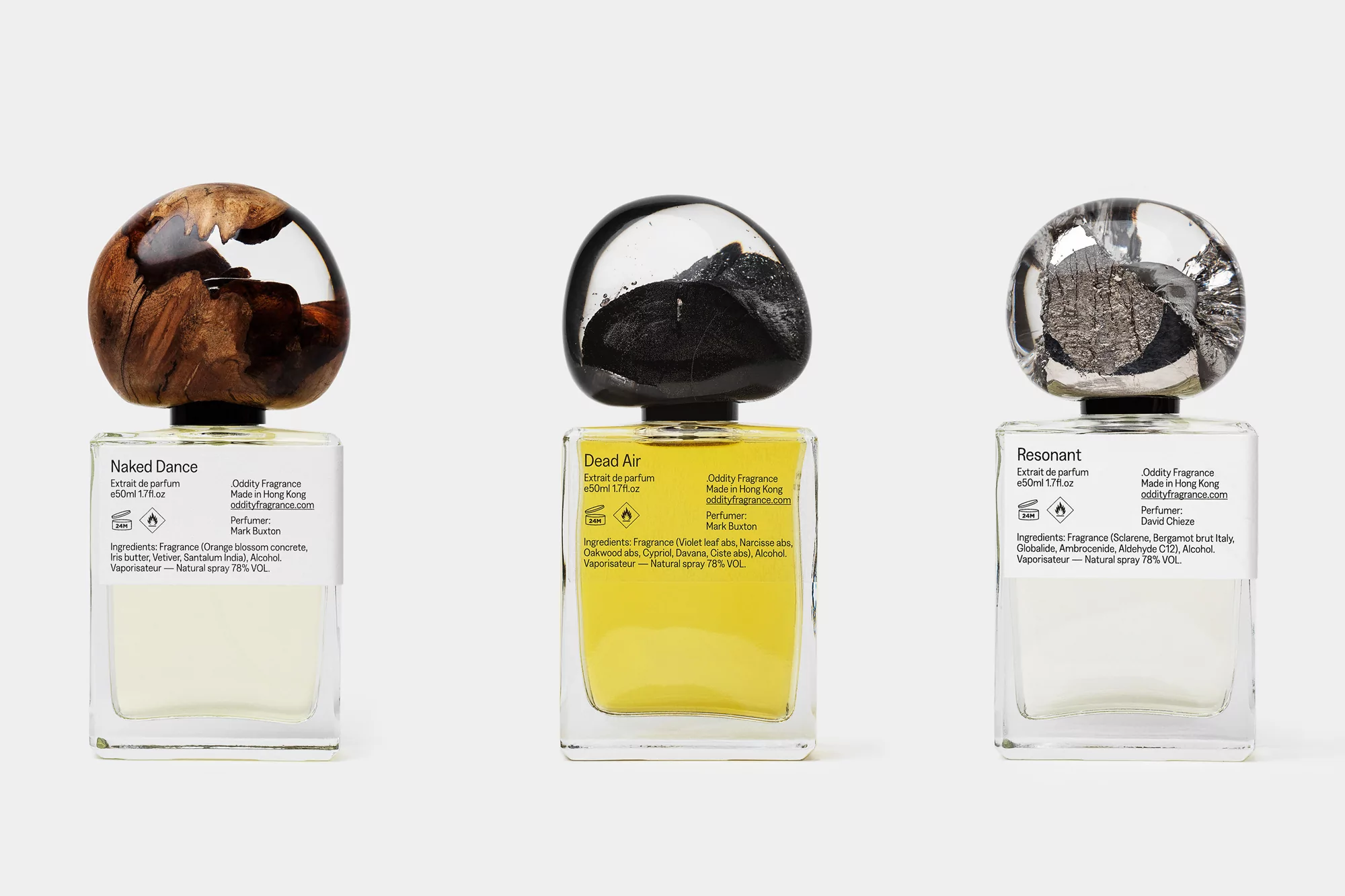 Three "Oddity Fragrance" perfume bottles lined up on a white surface, each with unique caps: wood, black stone, and silver.