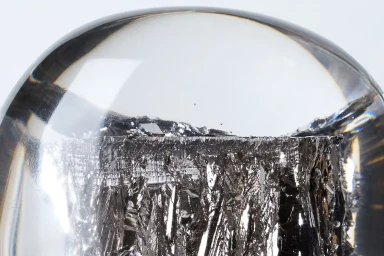 Close-up macro shot of a metallic crystal structure encased inside a clear glass dome against a plain white background.