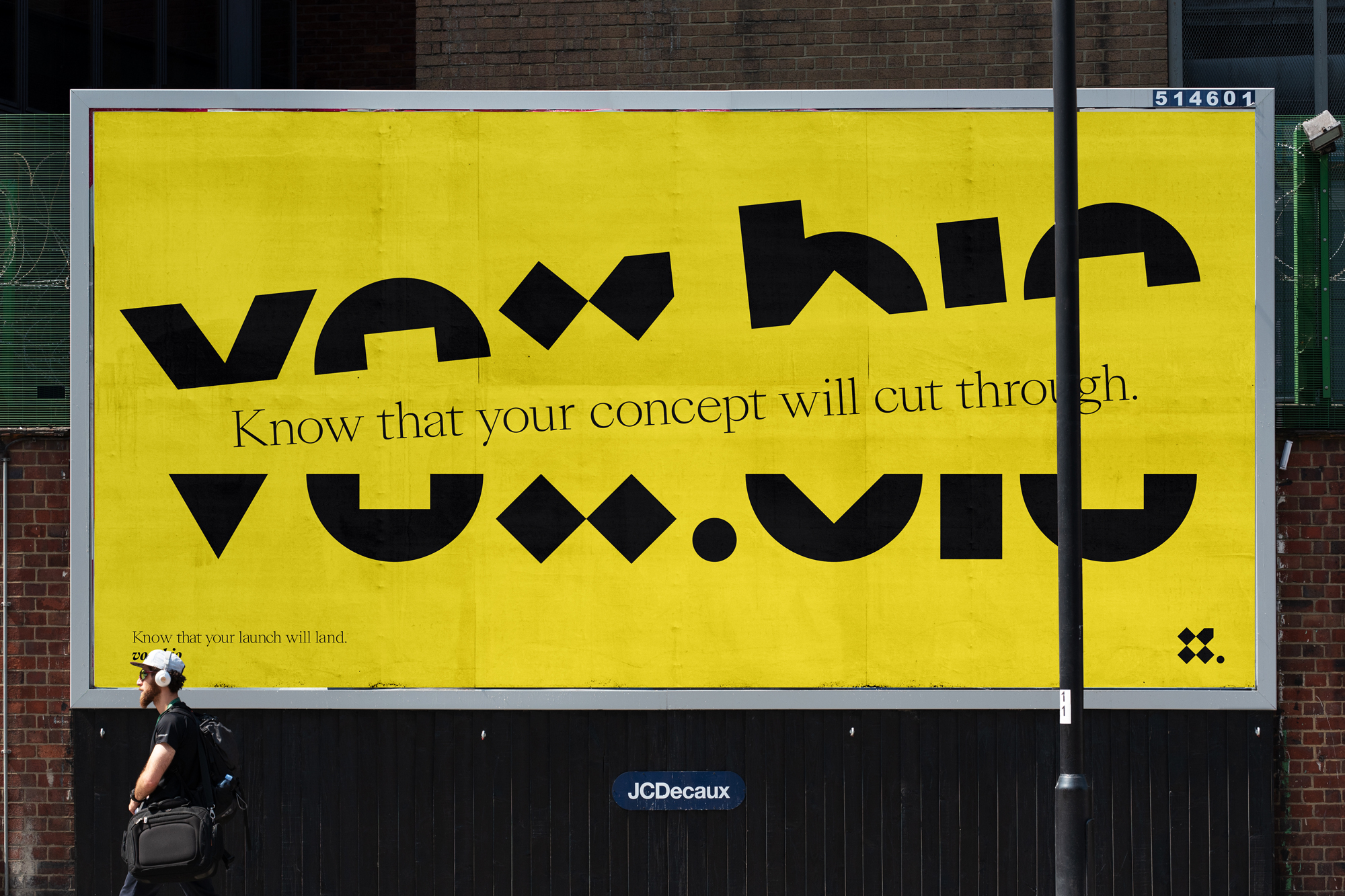 Eye-level view of a large yellow billboard with black geometric typography mounted on a brick wall with a man walking in the foreground.