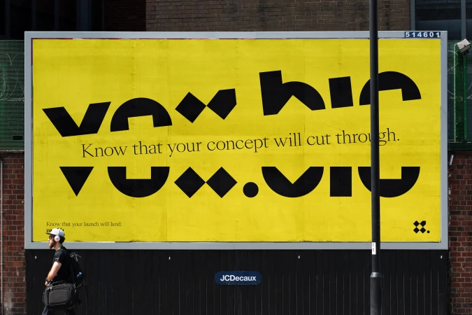 Eye-level view of a large yellow billboard with black geometric typography mounted on a brick wall with a man walking in the foreground.